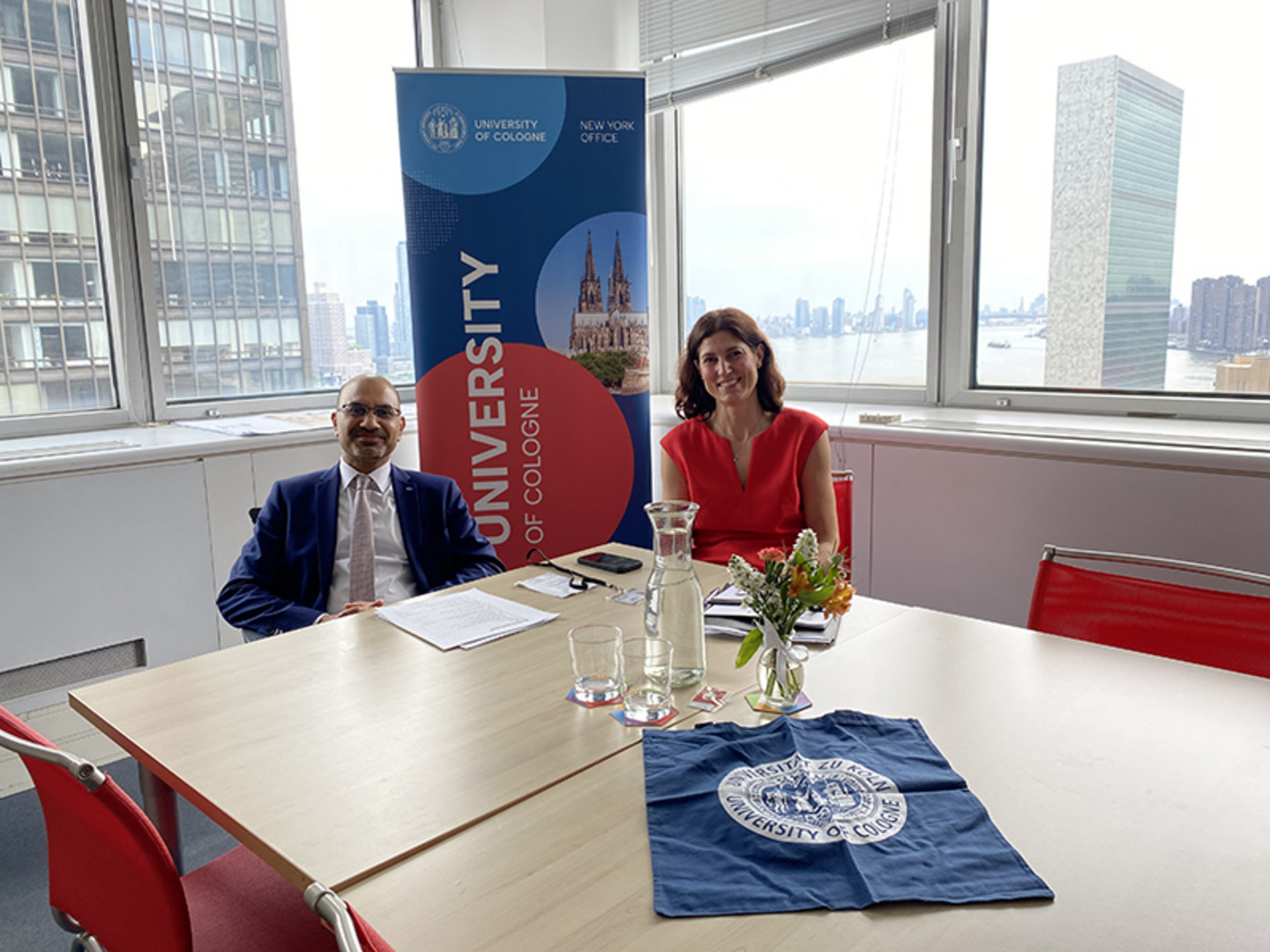 A man and a woman sitting at a table smile at the camera. In the background is the logo of the University of Cologne and the New York skyline.