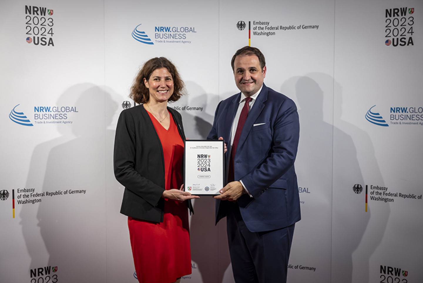 A woman and a man stand in front of a photo wall with the logos of the NRW-USA Year 2023/2024, the German Embassy in Washington and NRW Global Business. The man hands the woman a framed certificate on which the NRW-USA Year logo is also recognisable.