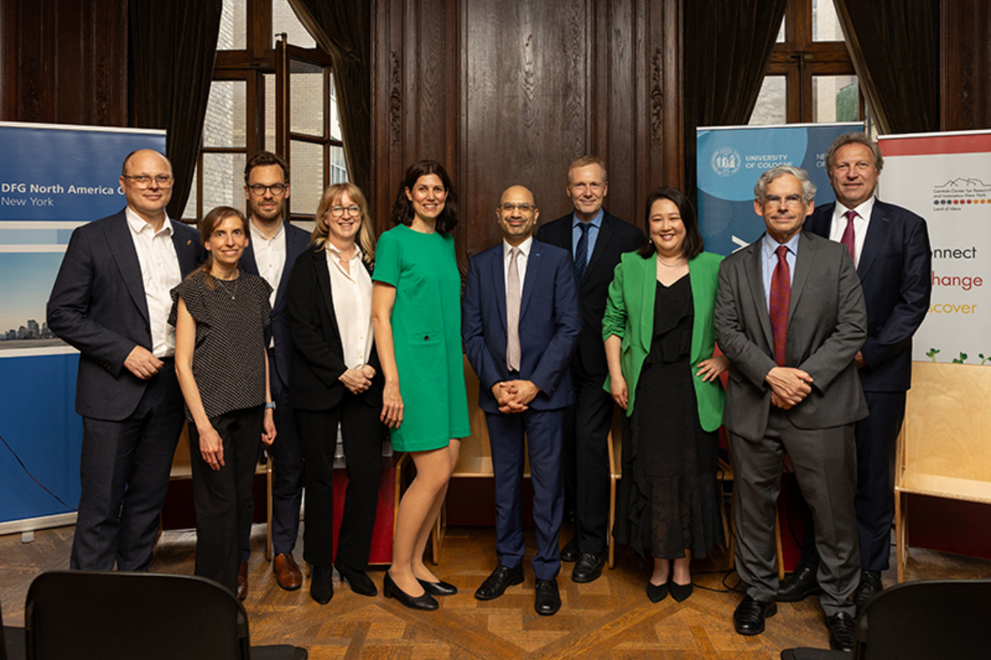 A group of ten people standing next to each other on a stage and looking into the camera. In the background are logos from the DFG, the University of Cologne and the DWIH.