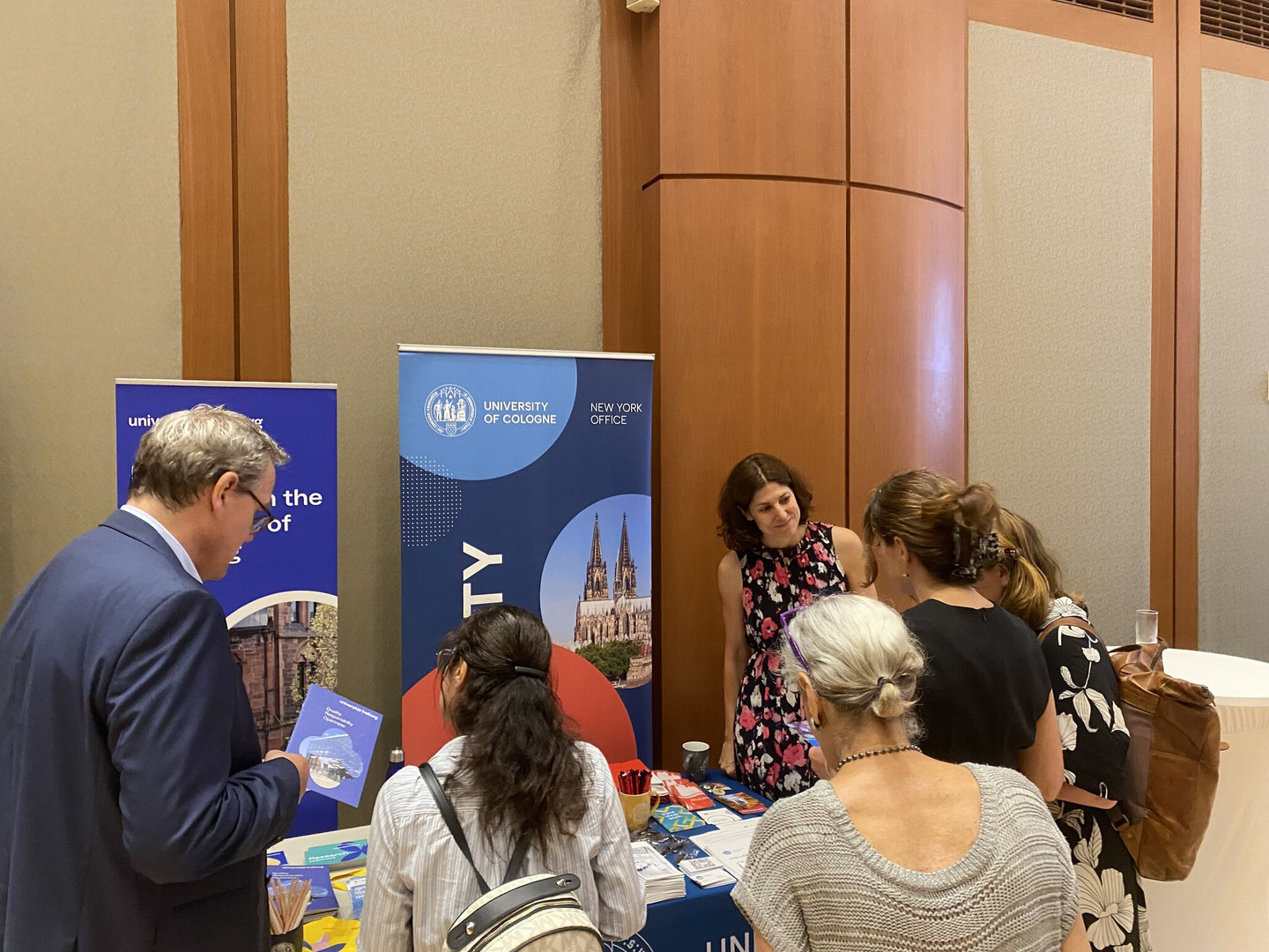 Six people are standing in a semi-circle around a table with information material and engrossed in conversation. The logo of the University of Cologne can be seen in the background.