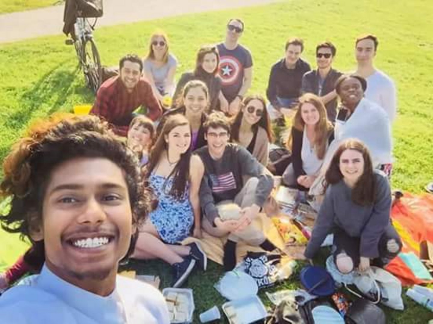 Selfie of 15 Students having a picnic in a park. They are sitting on blankets and have some food surronding them.