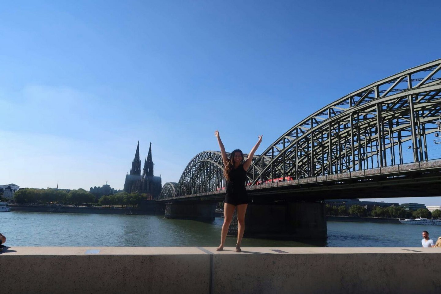Jaquelin Alonso standing on a wall in front of the river rhine, cologne cathedral and the hohenzollern bridge.