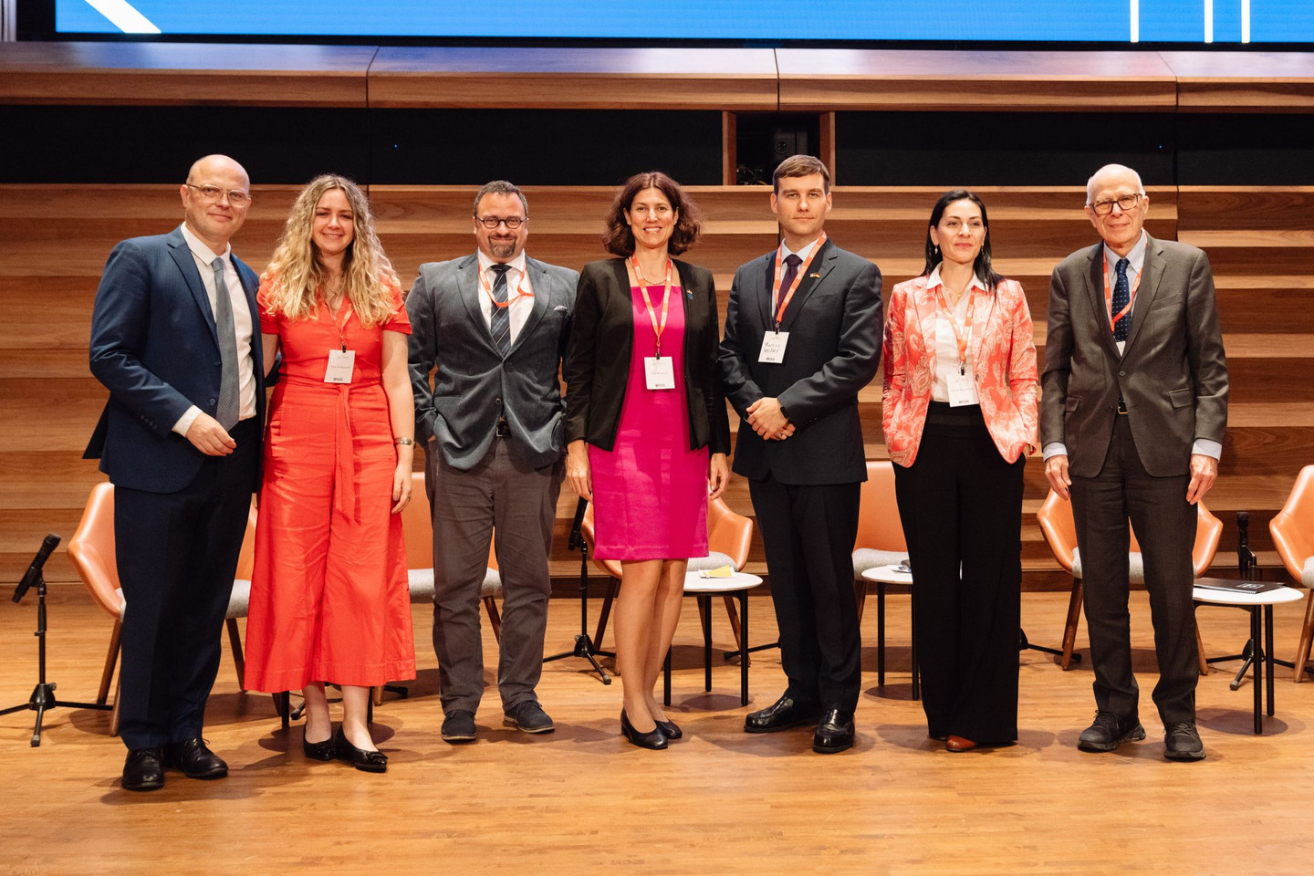 Seven people stand next to each other on a large stage and look smilingly into the camera.